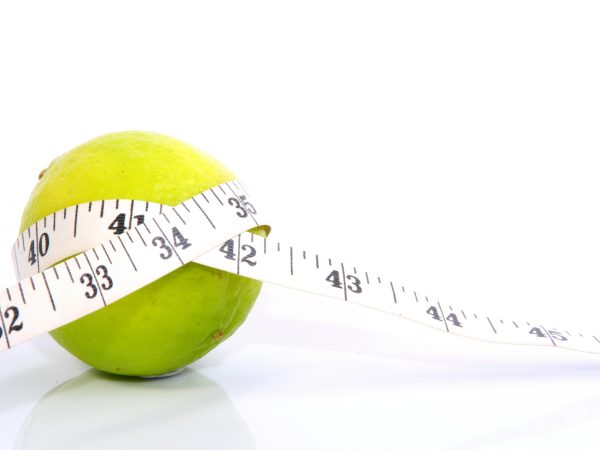 Grün-gelbe Zitrusfrucht auf weißem Hintergrund, mit Maßband umwickelt, als Sinnbild für Taillenumfangmessung, gesunde Ernährung und Gewichtsoptimierung.